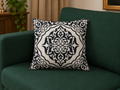 A square throw pillow with a black and white ornate damask pattern, resting on a dark green sofa in a living room setting.