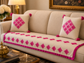 A beige sofa in a living room setting with a cream-colored slipcover featuring bright pink embroidered diamond patterns and matching pink pom-pom tassels. Two matching decorative throw pillows are placed on the sofa.