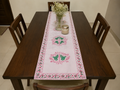 A white table runner with pink floral borders and green butterfly motifs, laid on a dark wood dining table with matching chairs and baby's breath flowers.