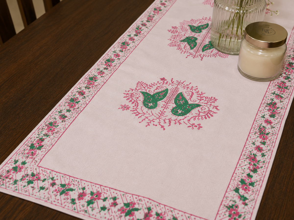 A rectangular table runner laid on a dark wooden table, featuring a pink floral border and two green embroidered motifs in the center. A glass jar candle and a ribbed glass vase sit on top.
