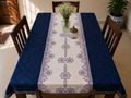 A navy blue tablecloth with a central cream table runner featuring purple floral and geometric patterns, styled on a wooden dining table with chairs and home decor.