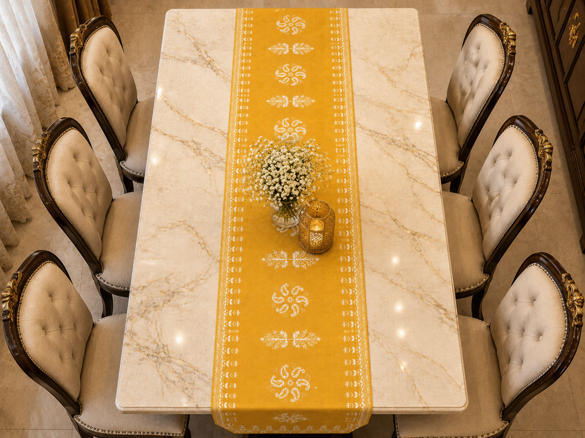 A dining table set with a long yellow table runner featuring white ornamental patterns, surrounded by six tufted cream chairs with dark wood frames. The table has a white marble-look surface and is decorated with a small floral centerpiece and a gold candle holder.