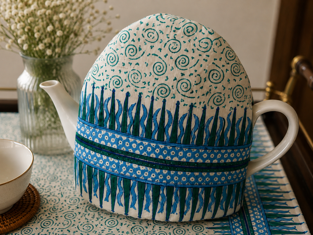 A white ceramic teapot covered with a decorative fabric sleeve featuring blue and green patterns, set on a patterned tablecloth with a small white cup and a vase of baby's breath in the background.