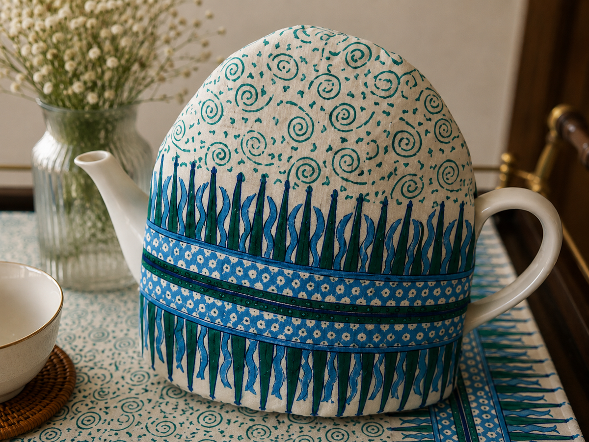 A white ceramic teapot covered with a decorative fabric sleeve featuring blue and green patterns, set on a patterned tablecloth with a small white cup and a vase of baby's breath in the background.