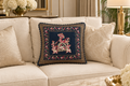 A decorative square throw pillow with a navy blue center and intricate multicolor embroidered figures, styled on a cream-colored sofa in a warm, elegant living room setting.