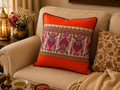 A square decorative throw pillow with a vibrant orange border and a central cream-colored panel featuring intricate magenta, green, and gold traditional patterns, styled on a beige sofa next to another patterned pillow.