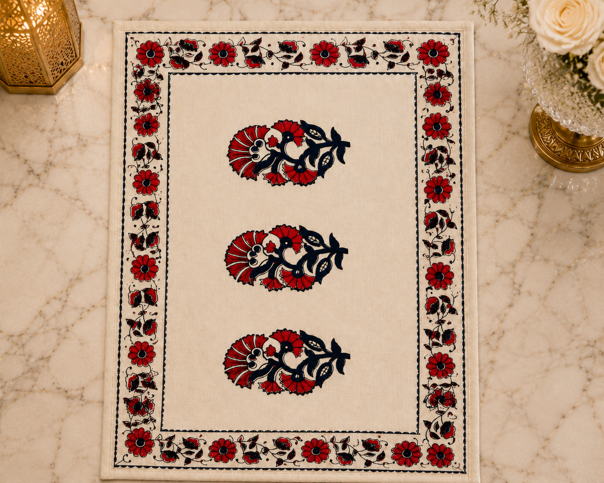 A rectangular cream-colored table mat with a black floral border and three central red and black floral motifs, displayed on a light marble surface with decorative props.