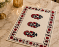 A rectangular cream-colored table mat with a black and red floral block print design, styled on a polished marble tabletop next to a gold lantern and a glass vase.